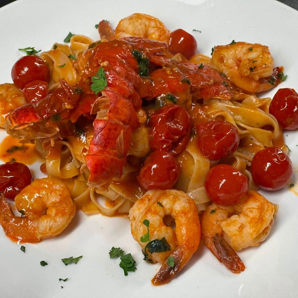 Tagliatelle topped with lobster, shrimp, cherry tomatoes, tomato sauce, and fresh parsley on a white plate.