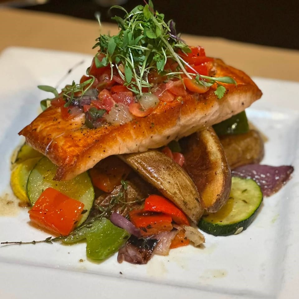 Seared salmon topped with diced tomatoes and microgreens, served with roasted potatoes and colorful grilled vegetables.