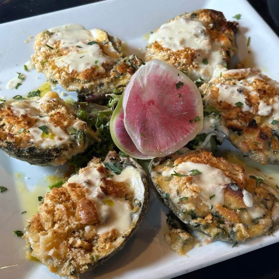 Five baked oysters topped with creamy sauce, toasted breadcrumbs, herbs, and radish slices on a white plate.