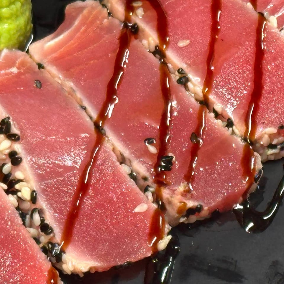 Sliced seared tuna coated with black and white sesame seeds, drizzled with sauce, served alongside wasabi.
