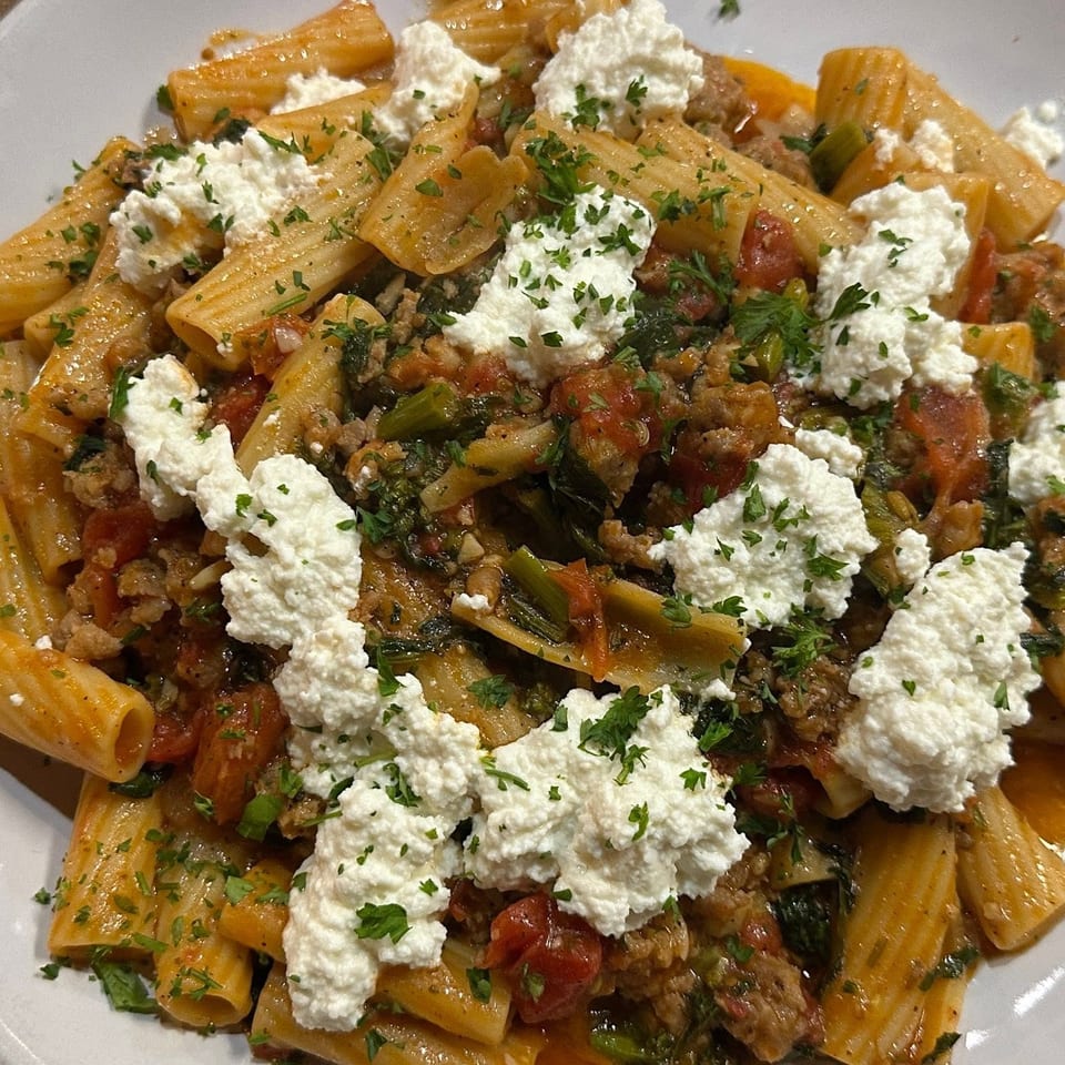 Rigatoni pasta with tomato sauce, sausage, vegetables, ricotta cheese, and chopped parsley on a white plate.