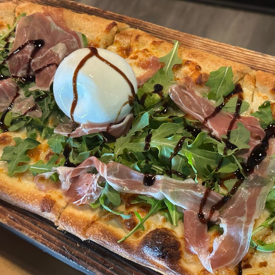 Flatbread topped with prosciutto, arugula, a ball of fresh cheese, and balsamic glaze on a wooden serving board.