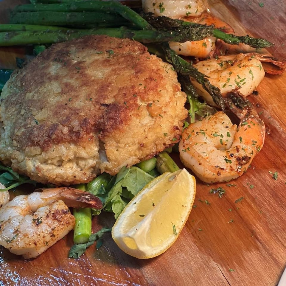 Golden-brown seafood cake with grilled shrimp, asparagus, and a lemon wedge on a wooden serving board.
