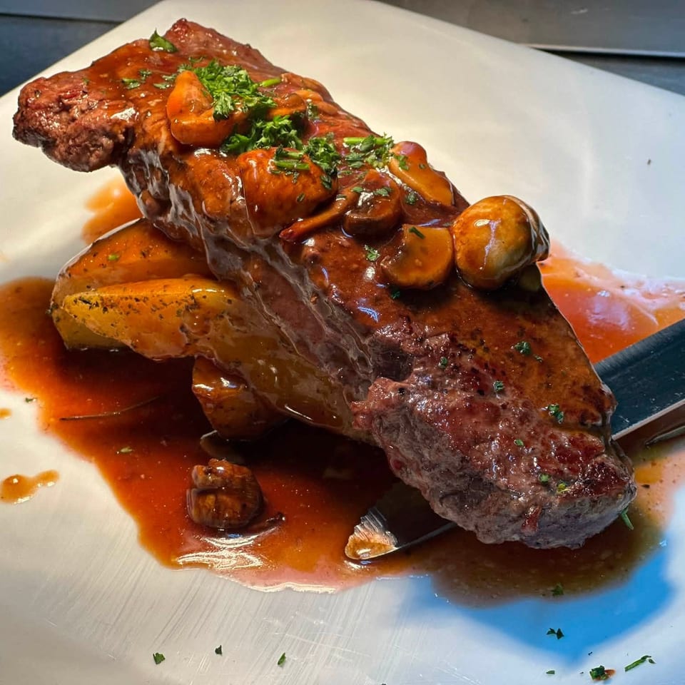 Grilled steak topped with mushrooms, carrots, herbs, and brown sauce, served on a white plate with roasted vegetables.